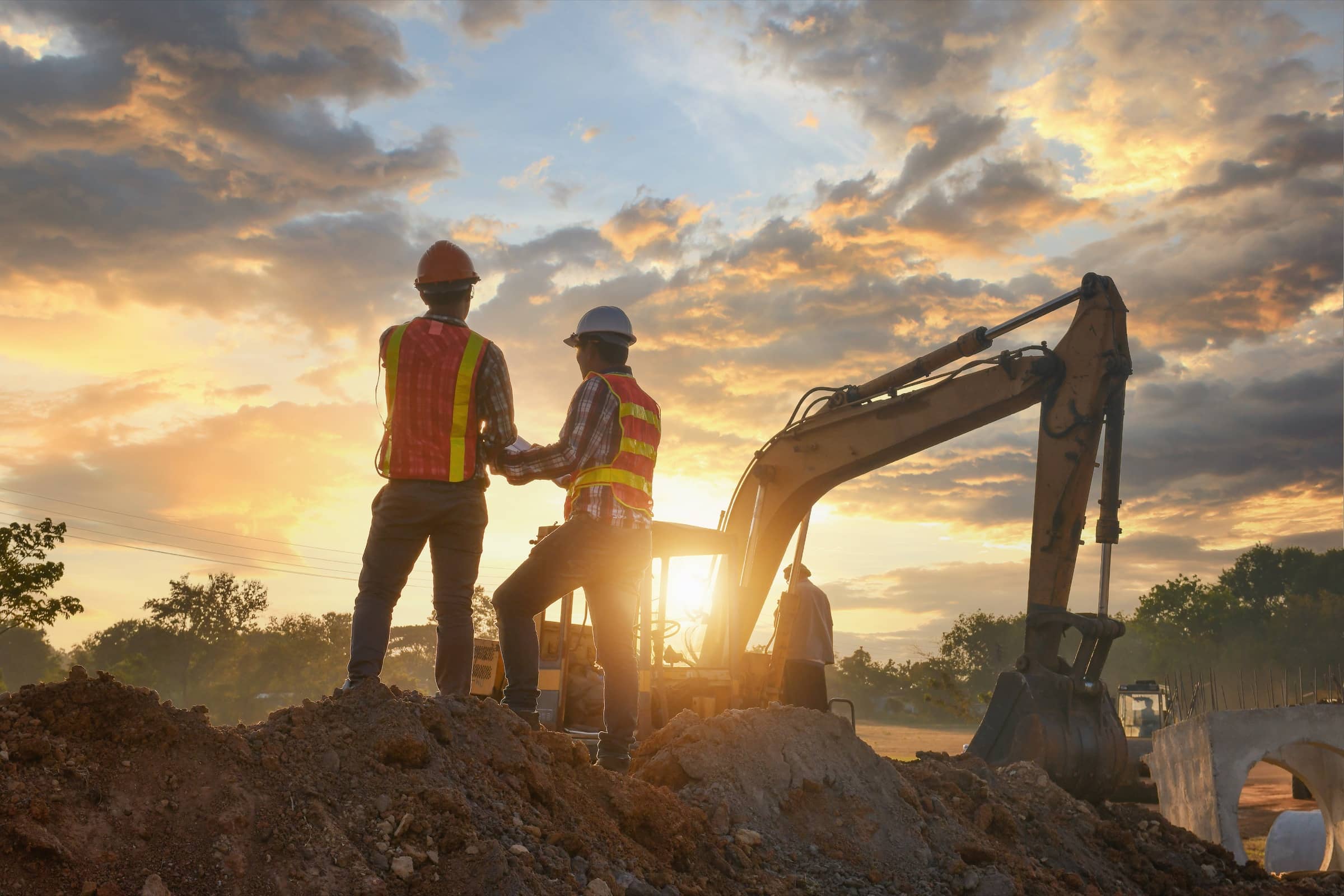 Two crew members in safety vests reviewing plans beside an excavator at sunset